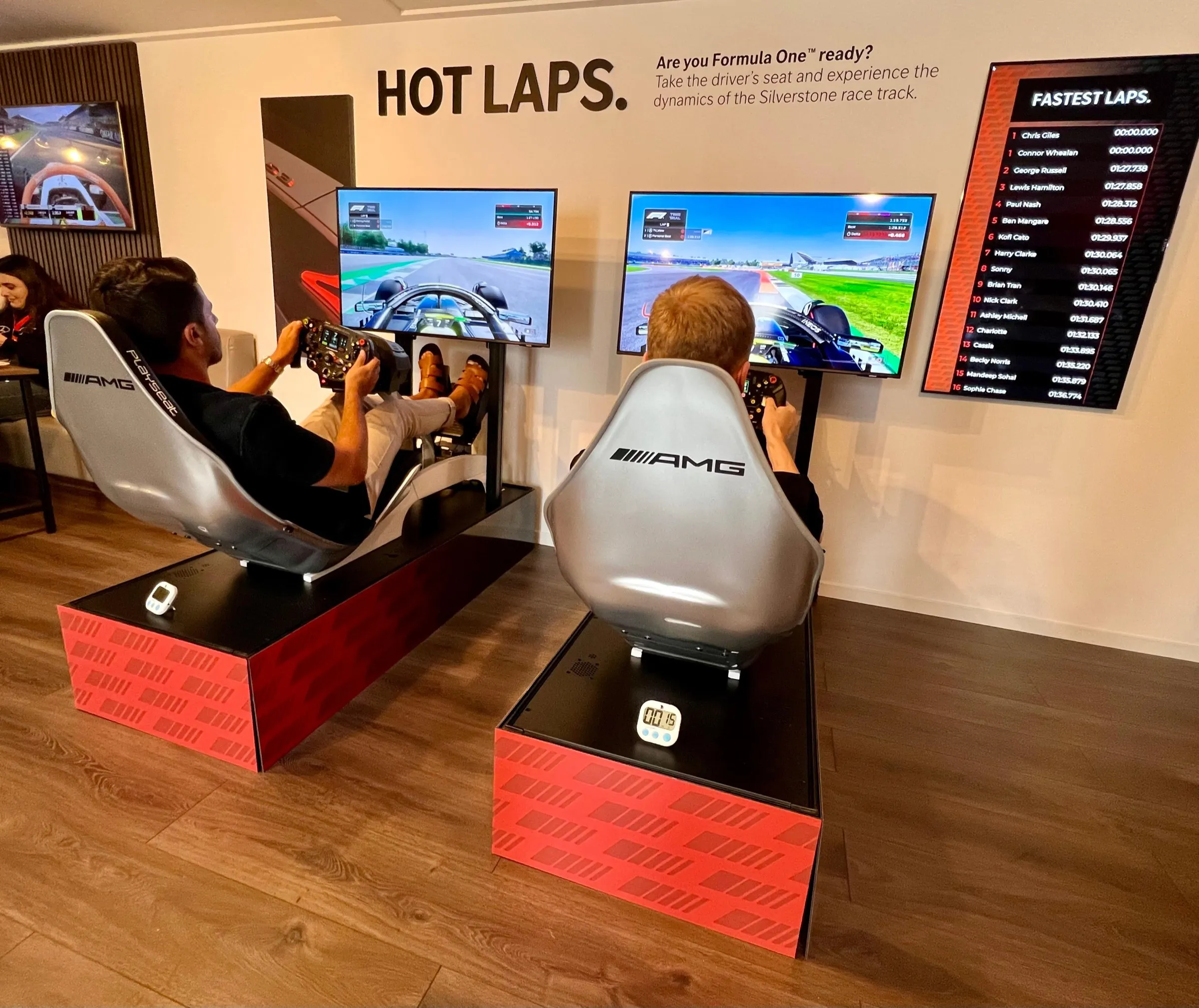 Two guests racing AMG branded simulators beside a wall mounted digital leaderboard listing the fastest laps