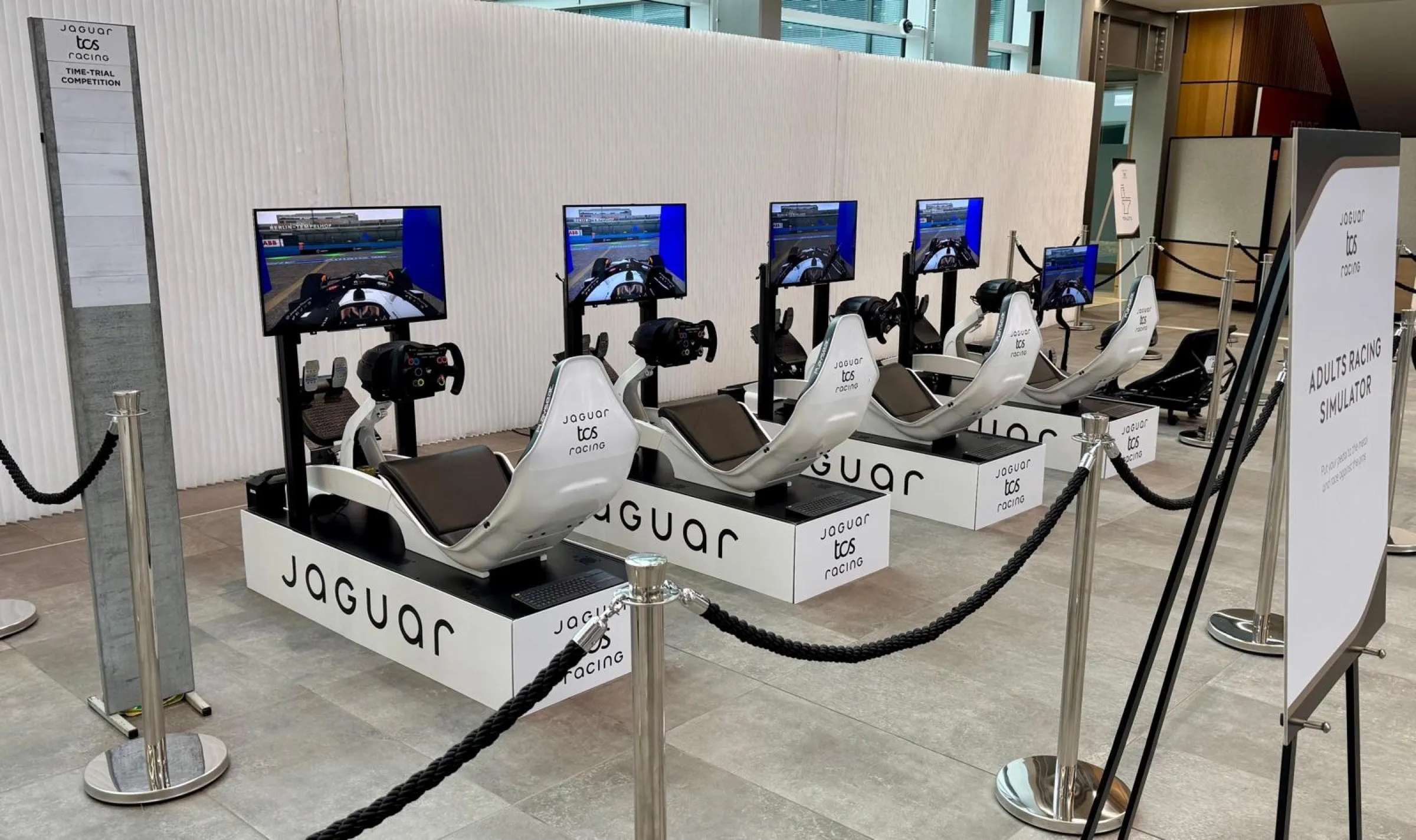 Row of white Jaguar TCS Racing simulators on branded plinths behind rope barriers, beside a sign reading Adults Racing Simulator