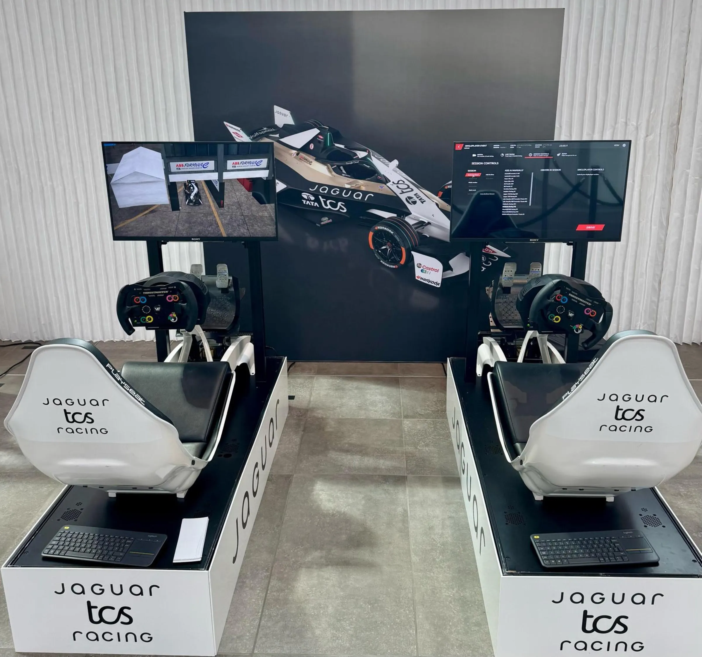 Two Jaguar TCS Racing branded simulators in front of a large image of the team Formula E car, with the racing game on the screens