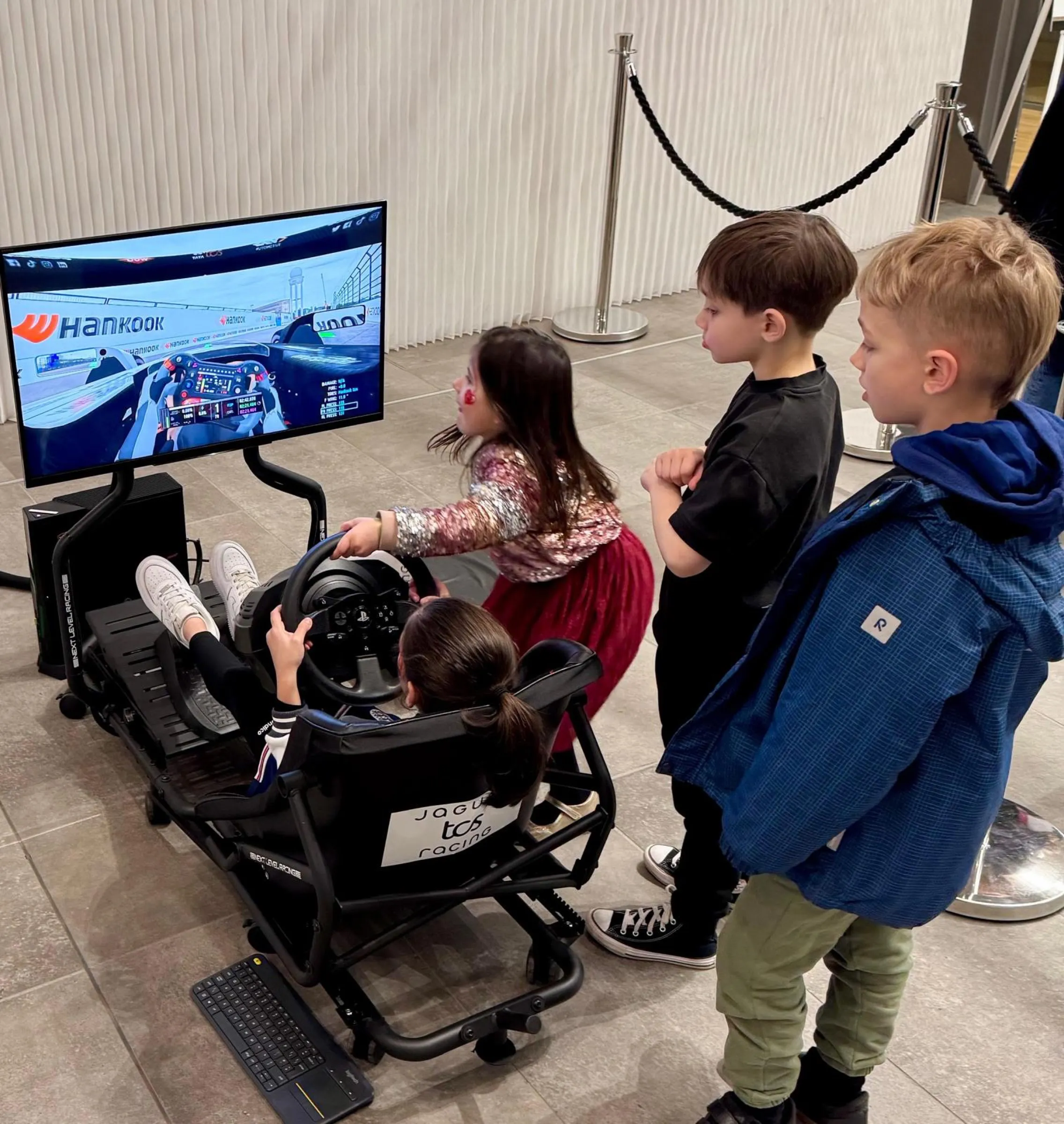 Child driving a Jaguar TCS Racing branded simulator with three other children watching the racing on screen