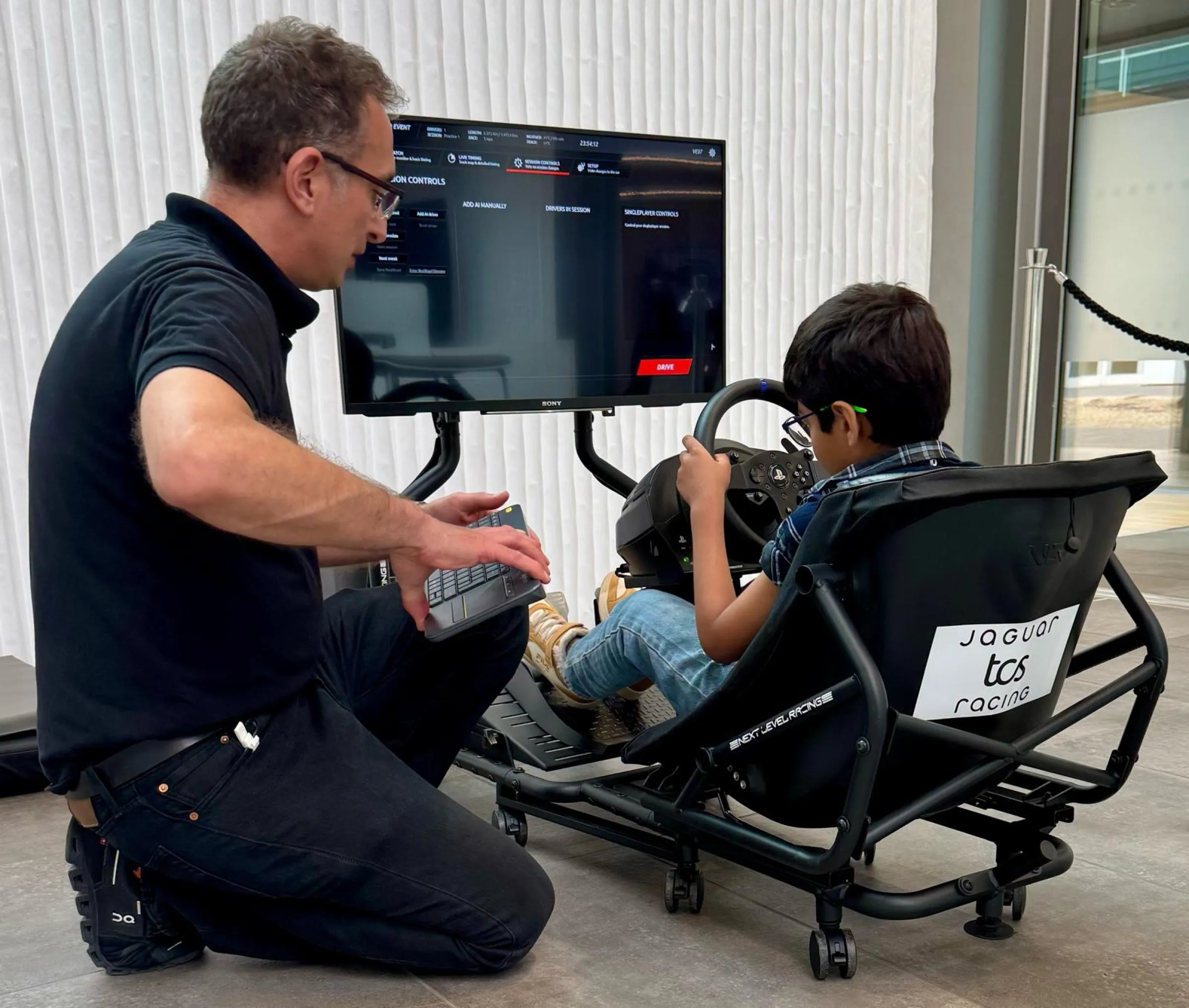 Virtually Entertained crew member kneeling beside a child in a Jaguar TCS Racing branded simulator, setting up the session on a keyboard