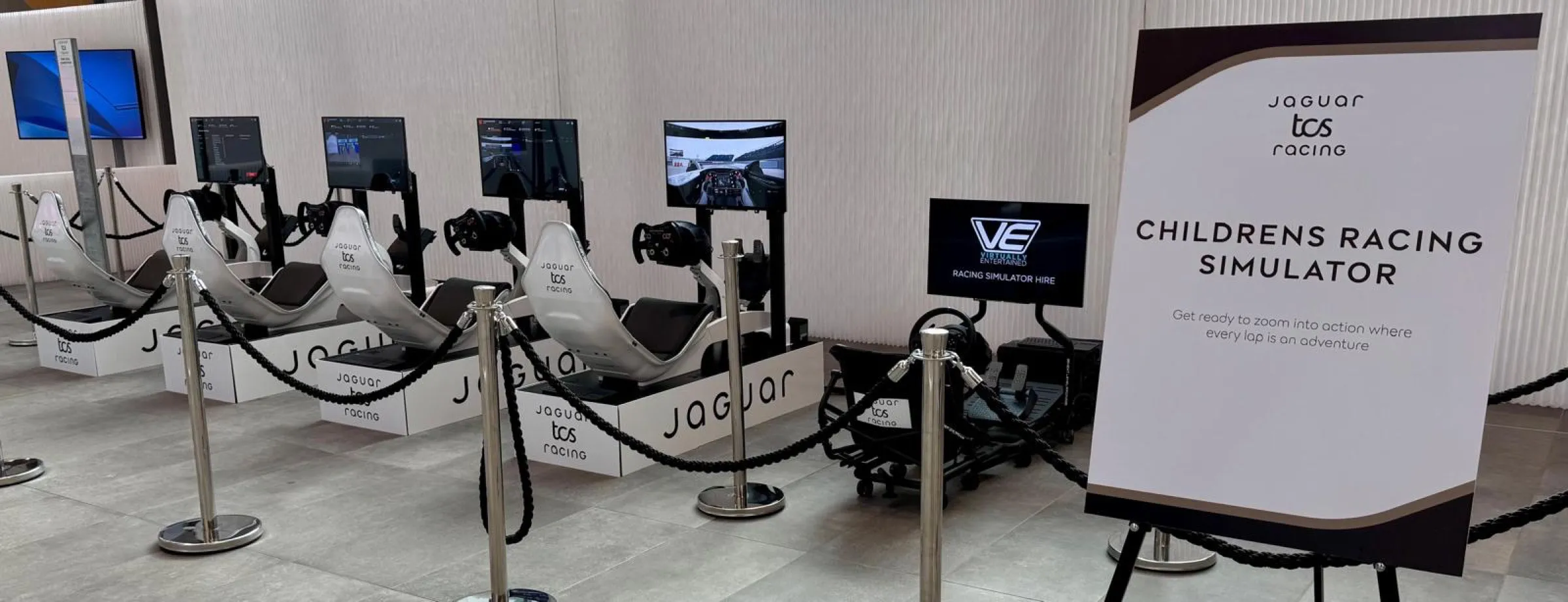 Row of white Jaguar TCS Racing branded simulators behind rope barriers next to a sign reading Childrens Racing Simulator