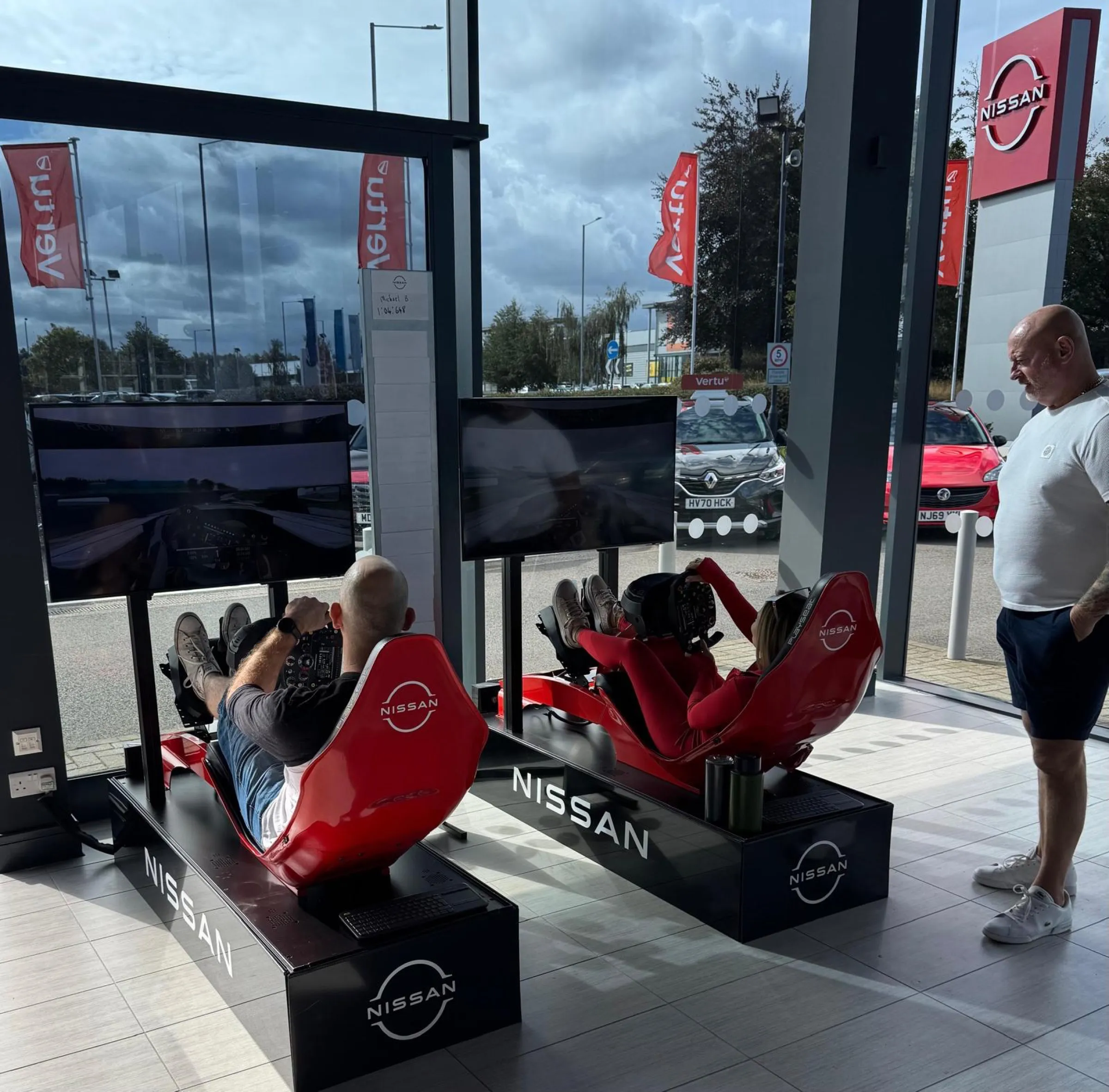 Guests racing two red Nissan-branded simulators inside a dealership showroom, with a printed leaderboard on the glass behind them
