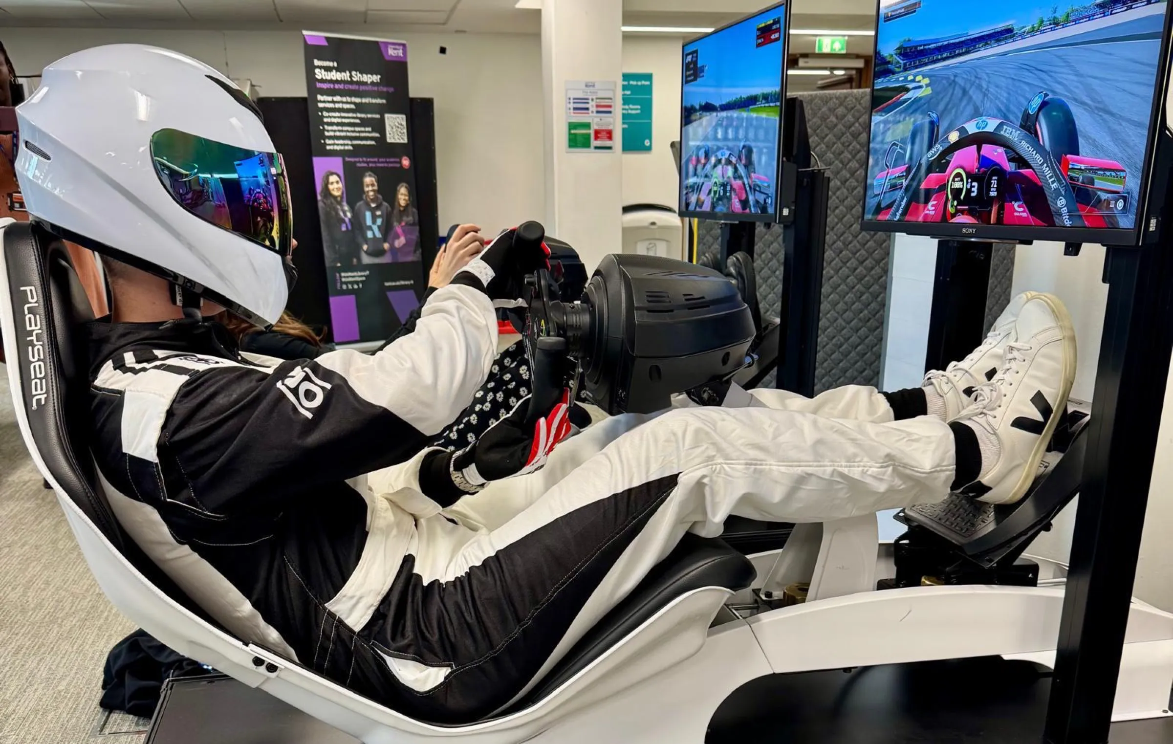 Driver in a race suit and helmet at the wheel of a simulator during a campus event